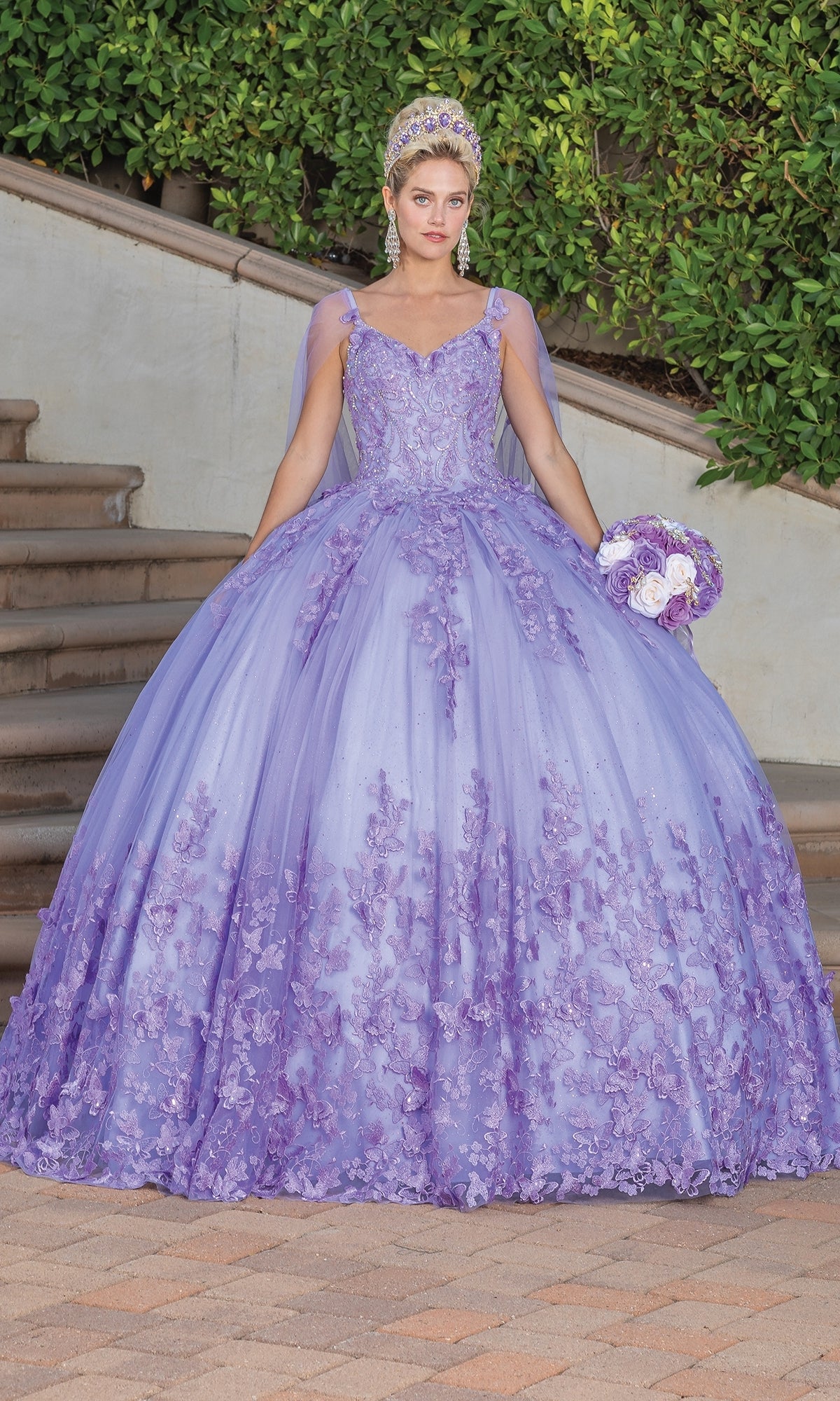 A woman in the Quinceanera Dress 1746 By Dancing Queen—a lavender A-line ball gown with floral embroidery and a sweetheart neckline—stands outdoors with a bouquet, surrounded by greenery and stone steps.